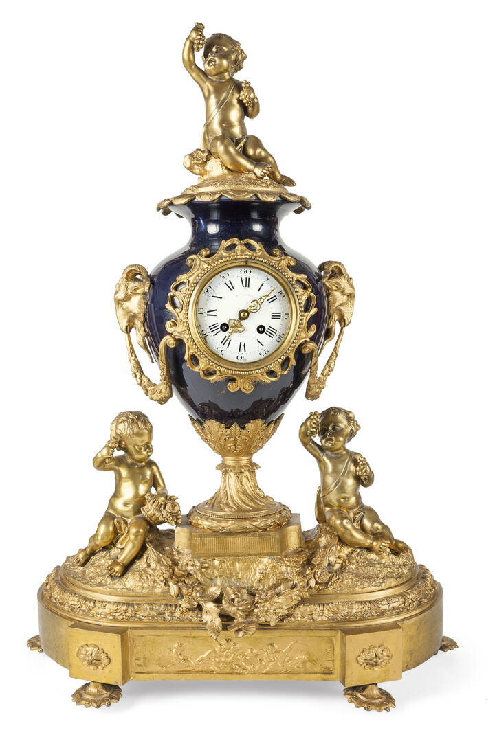 OROLOGIO DA TAVOLO - Orologio in bronzo dorato e porcellana decorato a putti e teste di caprone, fine XIX. Firmato sul quadrante ' F.lli Rier Paris'. In parte ridorato<br>H cm 74